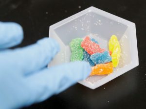 A blue-gloved hand holding a hexagonal mold containing sugar-coated, colorful MDMA gummy bears.