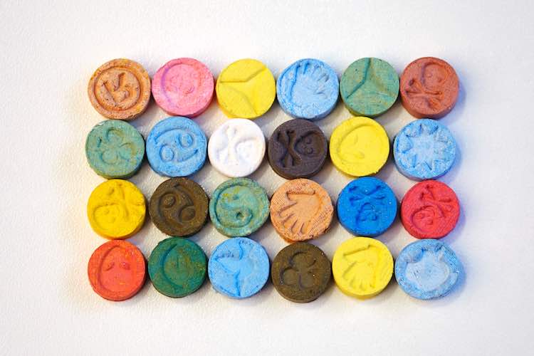 Grid of colorful ecstasy tablets showing wide variation in colors (pink, yellow, blue, green, red, orange, brown) and imprinted logos/symbols. Illustrates how MDMA pills can look vastly different—no reliable way to judge contents by appearance alone.