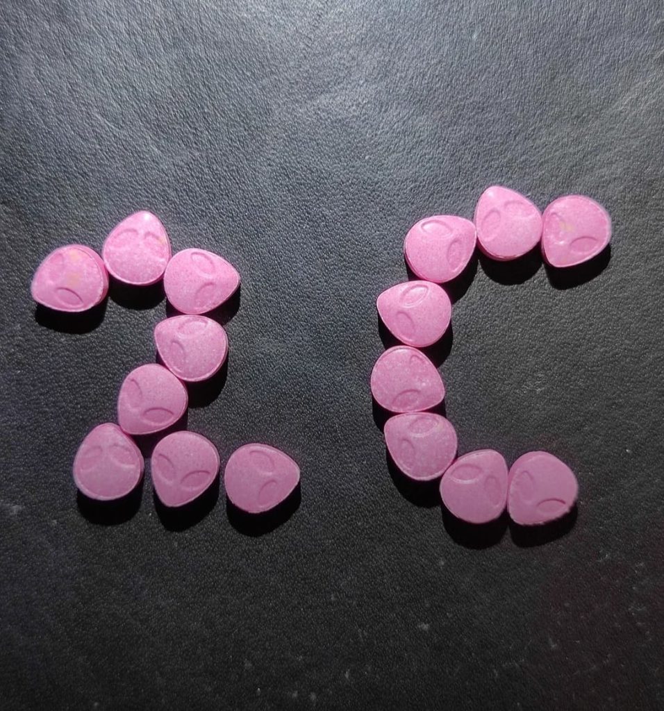 Pink 2C-B Pills with the compressed Heart Alien logo arranged to spell the letters 2C on a dark surface.